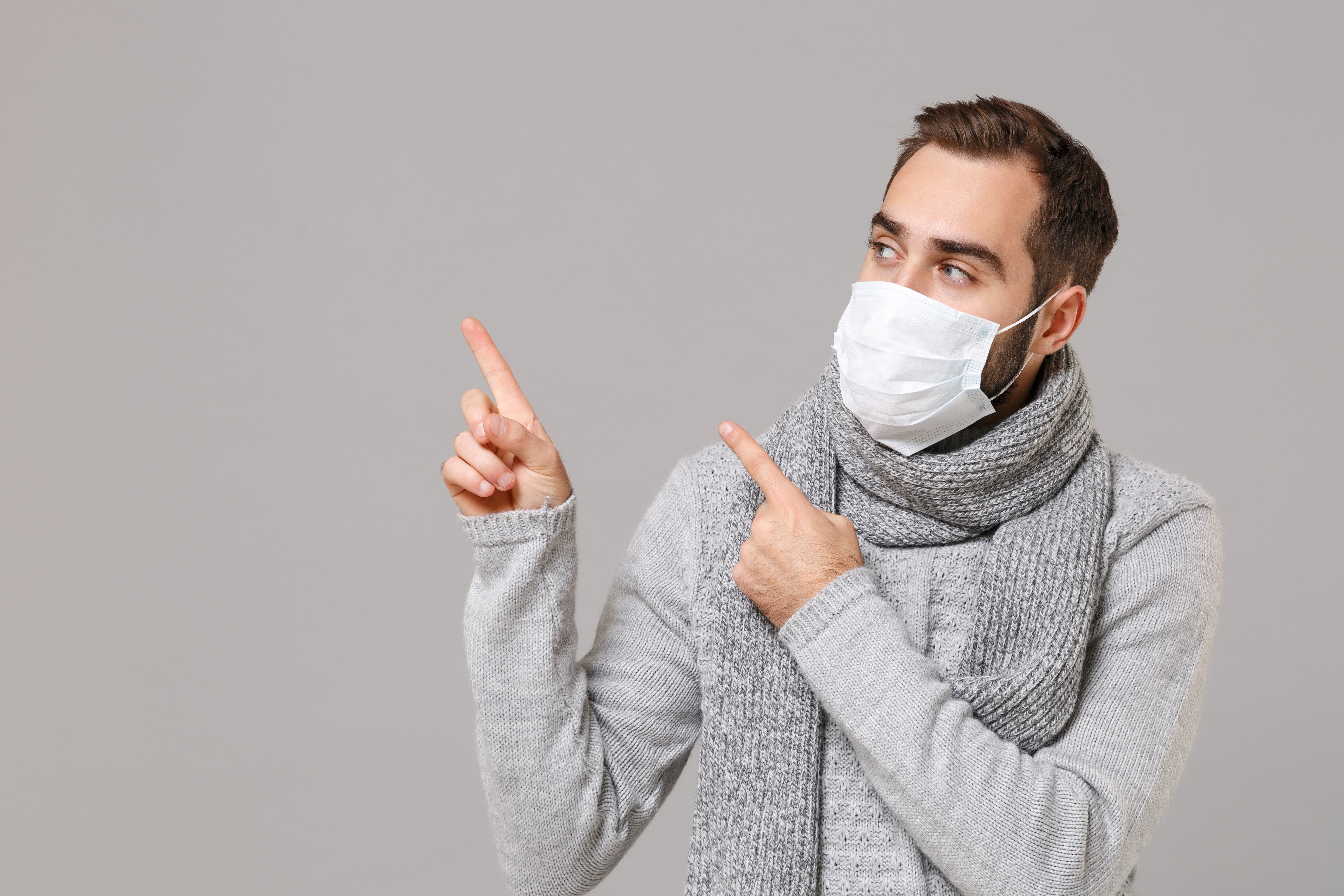 Young man in gray sweater, scarf isolated on grey background. Healthy lifestyle, ill disease treatment, cold season concept. Mock up copy space. Wearing sterile face mask point index fingers aside up