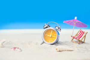 Summer hours – an alarm clock sitting on a sandy beach with an umbrella and chair.